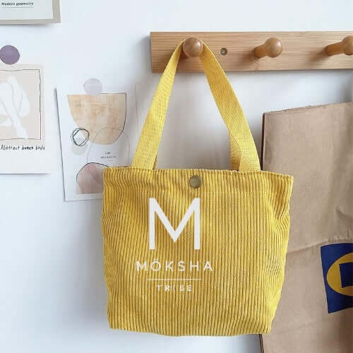 Möksha Tribe Tote Bag in yellow, displayed on a wall with accessories, showcasing its unique design and vibrant color.