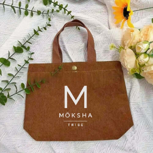 Möksha Tribe Tote Bag in brown with logo on a textured surface surrounded by flowers.