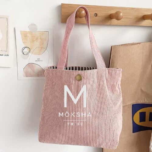 Möksha Tribe Tote Bag displayed on a hook with pink corduroy fabric and logo.