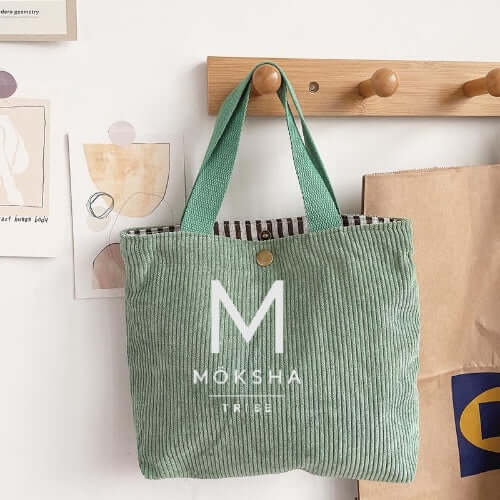Möksha Tribe Tote Bag in green fabric hanging on a hook, showcasing its logo and design.